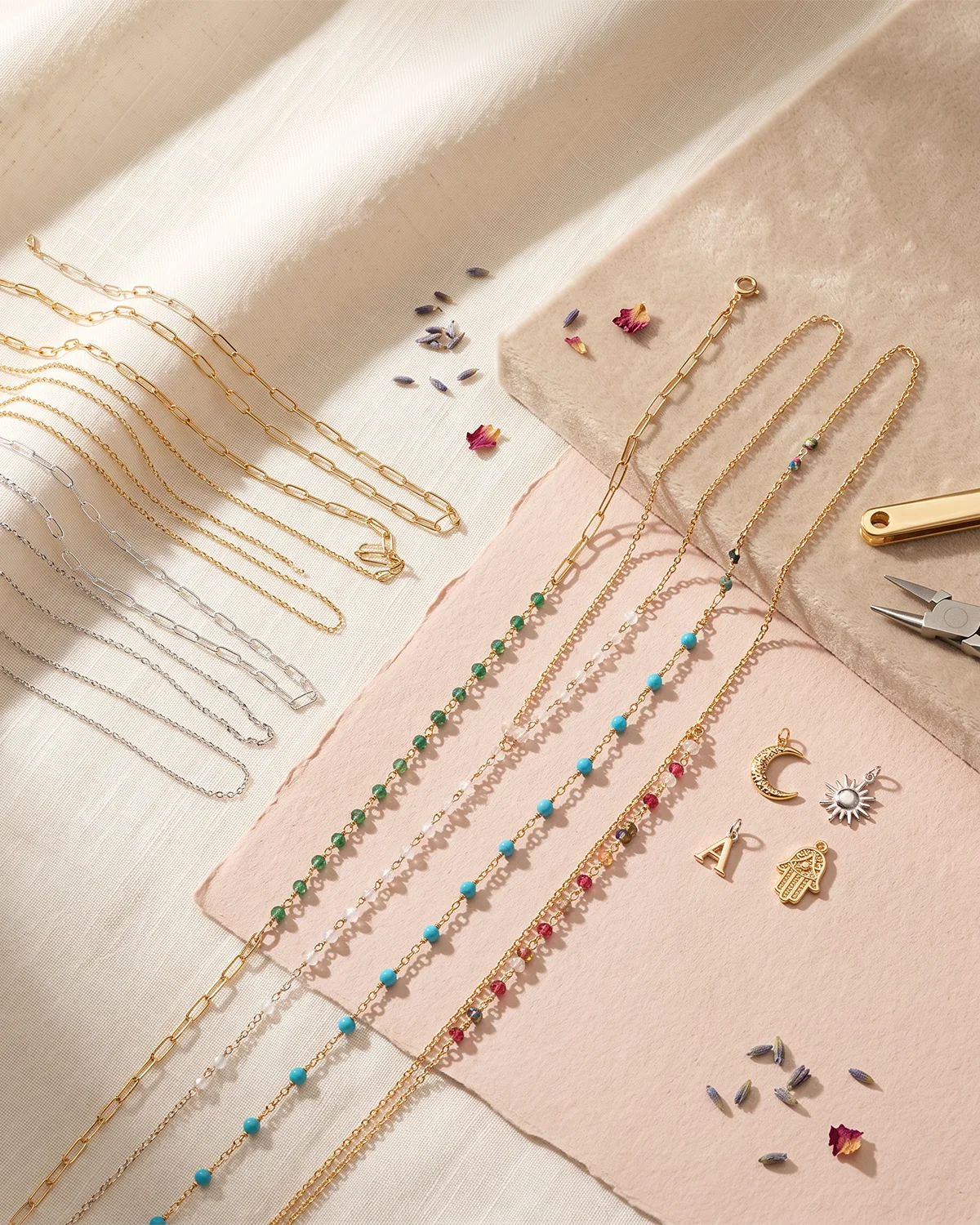 Fine jewelry chains and charms arranged across ivory, sand and blush surfaces