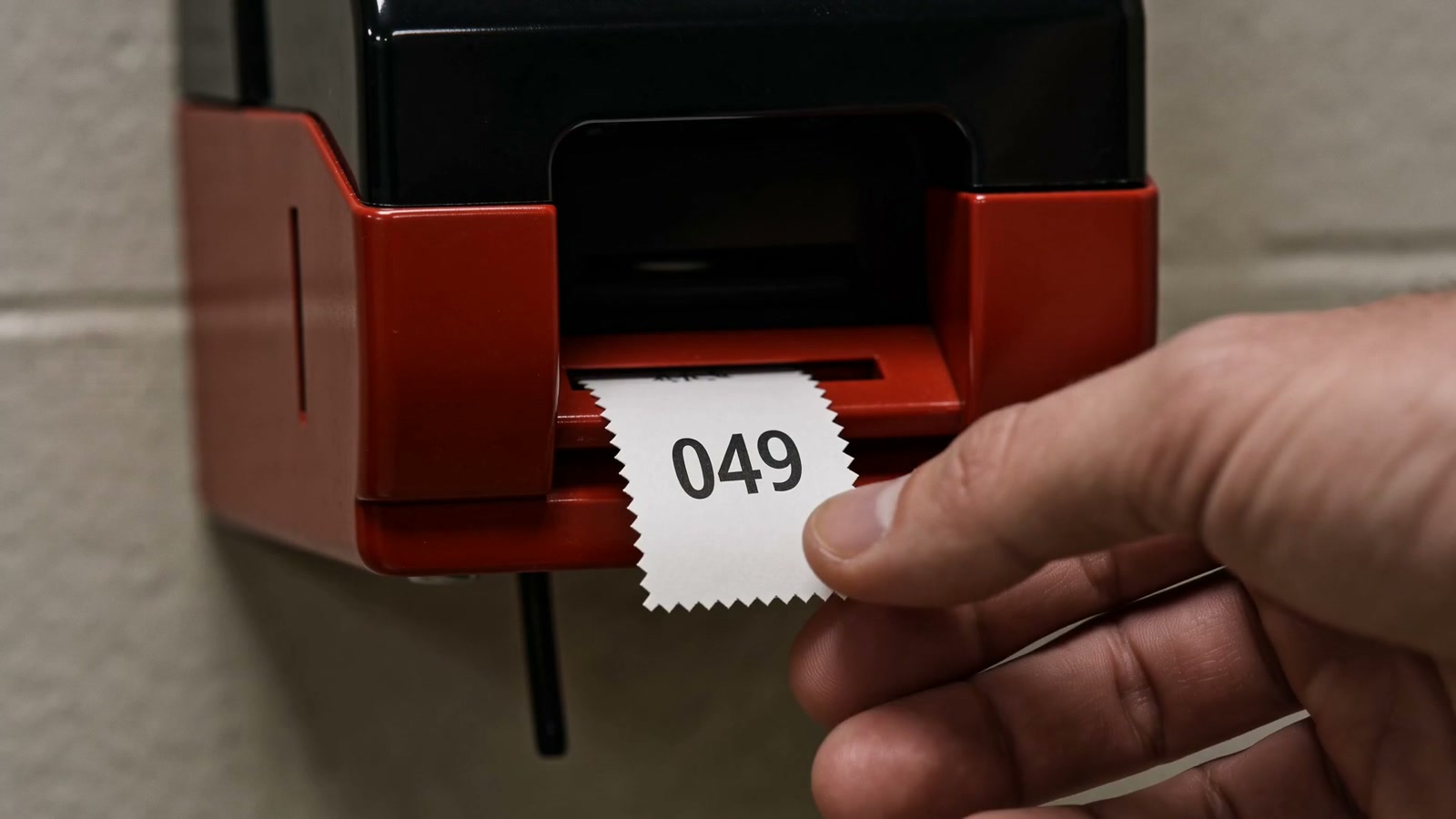A hand pulls ticket 049 from a red waiting room dispenser