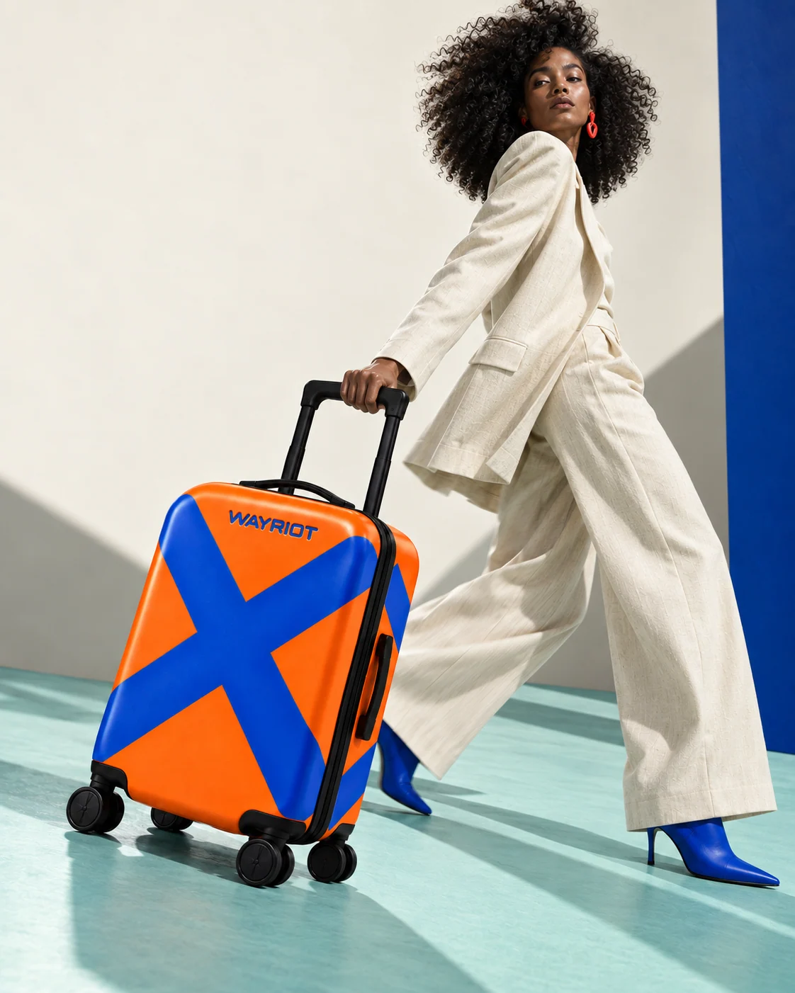Woman in ivory tailoring and cobalt boots walking with her orange Bold X suitcase